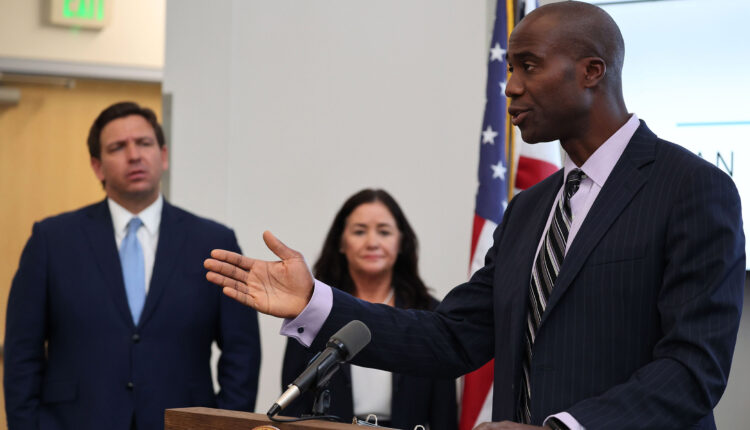 Florida Surgeon General Joe Ladapo speaks at a podium. Florida Gov. Ron Desantis stands beside him.