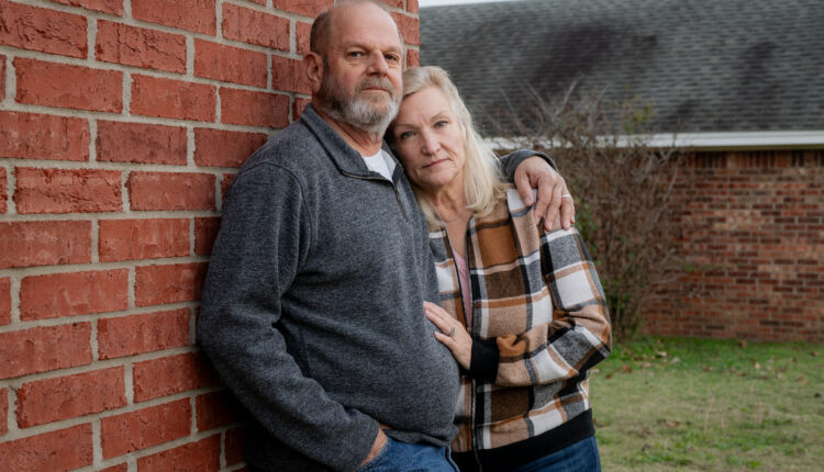 Ron Winters and his wife, Teresa, stand outside their home. Ron leans up against a brick wall, while his wife leans against him and places her hand on his stomach. They both look towards the camera.