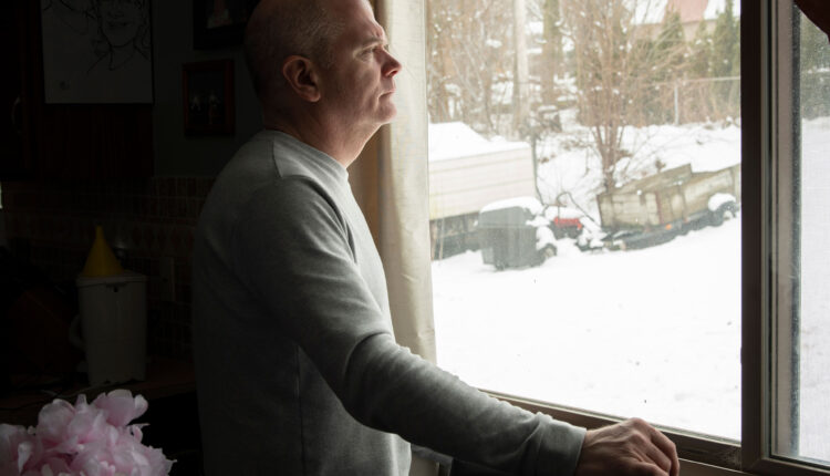 A side-view of a man standing at a window and looking out. His right had rests on the window sill.