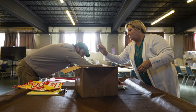 Two people in a warehouse, a woman in a white cardigan and a man in a green cap, sort syringes into a large brown box in a church basement.