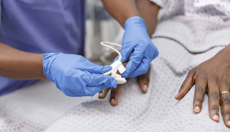 A close-up photo of a medical professional placing a pulse oximeter on the finger of a hospitalized patient who is lying in bed.