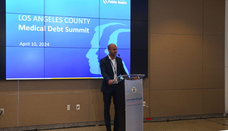 Naman Shah stands at a podium in a conference-like room. There is a projector screen behind him. A slide with a blue background and large white text reads, " Los Angeles County Medical Debt Summit, April 10, 2024."